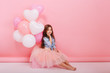 © Look! - Happy brightful image of cute joyful little girl in tulle skirt sitting on present with balloons isolated on pink background. Amazing charming birthday fashionable kid looking to camera