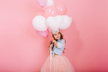 Wall Mural - Sweet lovely moments of happy childhood with flying balloons of amazing pretty young girl in tulle skirt smiling to side isolated on pink background. Charming happy kid celebrating birthday, carnival
