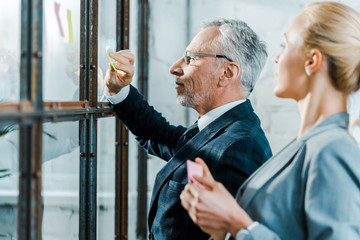 Wall Mural - selective focus of businessman in eye glasses putting sticky notes on window near businesswoman