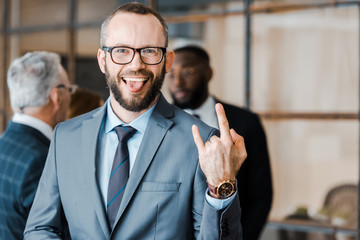 Wall Mural - selective focus of happy businessman showing tongue and rock sign near multicultural coworkers