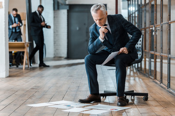 Wall Mural - pensive businessman sitting on chair and looking at charts and graphs near multicultural coworkers