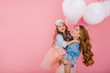© Look! - Adorable laughing birthday girl with colorful balloons embracing her young smiling mom after funny event. Attractive mother posing with pretty daughter in mask at party isolated on pink background