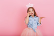 © Look! - Joyful little girl with long brunette hair in tulle skirt holding princess crown on head isolated on pink background. Celebrating brightful carnival for kids, expressing positivity of birthday party