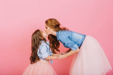 Wall Mural - Stylish curly mom and daughter hold hands and kiss sweetly at children's event on the pink background. Little long-haired girl in denim shirt and lush skirt kissing her young mother at birthday party
