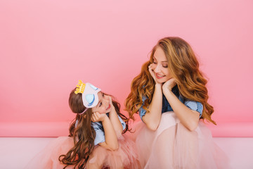 Beautiful young woman with trendy hairstyle sitting on the floor next to charming little girl and looking at her with love. Portrait of adorable curly mother having fun with child at birthday party