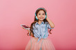 © Look! - Expressing positivity of happy child listening to music through blue headphones isolated on pink background. Pretty little girl with long brunette hair smiling to camera in tulle skirt