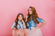 © Look! - Cheerful young mother and daughter fooling around wearing crown and carnival mask at family party. Two charming sisters in same vintage outfit playing together and smiling on pink background