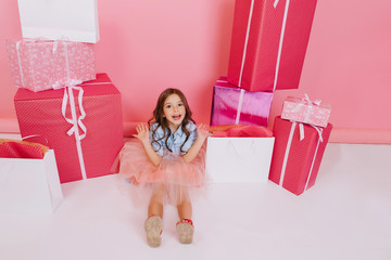 Wall Mural - View from above expressing to camera of sweet little girl in tulle skirt sitting on white floor suround colorful presents, giftboxes on pink background. Happy childhood, enjoying birthday