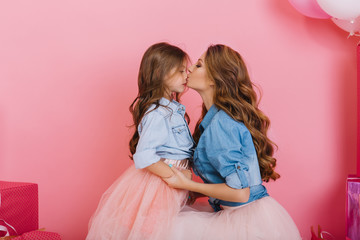 Wall Mural - Attractive young woman kisses her daughter in the nose holding her hands on pink background. Portrait of little birthday girl cute posing with curly stylish mom at the party with gifts and balloons
