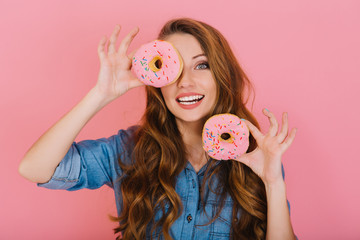 Long-haired adorable girl in denim shirt plays with glazed donuts before tea-drinking with friends. Excited charming young woman with curly hair happy to buy her favorite sweet doughnuts in bakery.