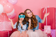 © Look! - Two adorable sisters wearing funny colorful carnival masks sitting next to present boxes. Family portrait of charming young mother and cute lovely daughter posing after birthday party with gifts