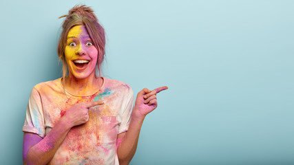 Horizontal studio shot of happy European woman points at side, covered with Holi colors, wears dirty t shirt, attracts your attention at blank space over blue background, laughs from positive emotions