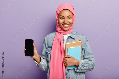 Satisfied Charming Young Muslim Woman In Pink Hijab On Head Holds Modern New Smartphone Device With Mockup Screen For Your Information Carries Spiral Pocketbook And Notepad Smiles Broadly Stock Photo Adobe