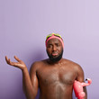 © WHstudio Leushin N - Upset black man gestures with puzzlement, raises hand, upset as can not swim well, has water drops on skin, carries inflated flamingo which helps not to drown, wears protective goggles for diving