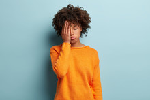 Photo Of Tired Dark Skinned Female Covers Face, Closes Eyes, Feels Fatigue, Needs Good Rest, Dressed In Orange Jumper, Has Sleepy Expression, Isolated Over Blue Background. Weariness And People
