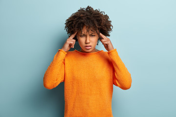Wall Mural - Waist up shot of distressed unhappy dark skinned woman touches temples, frowns face from dissatisfaction, suffers from headache or migraine, wears orange jumper, isolated on blue background.