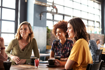 Three Young Female Friends Meeting Sit At Table In Coffee Shop And Talk