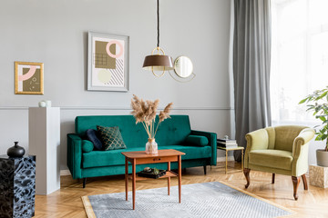 Stylish and elegant living room of apartment interior with green velvet armchair and sofa, brwon table, plant, design lamp and chic accessories. Abstract paintings on the gray wall. Luxury home decor.