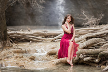 Young Beautiful Woman Is Wearing Fashion Red Dress Sitting In Forest Near The Waterfall