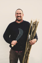Bearded Man Holding Bill Hook And Bunch Of Wooden Stakes For Traditional Hedge Laying, Smiling At Camera.