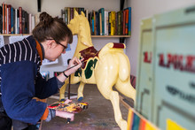 Woman Standing In Workshop, Painting A Traditional Wooden Carousel Horse From Merry-go-round.,Signwriter