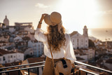 Fototapeta Psy - Blonde woman standing on the balcony and looking at coast view of the southern european city with sea during the sunset, wearing hat, cork bag, safari shorts and white shirt