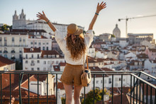 Blonde Woman Standing On The Balcony And Looking At Coast View Of The Southern European City With Sea During The Sunset, Wearing Hat, Cork Bag, Safari Shorts And White Shirt