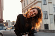 © alones - Funny positive young woman in stylish black clothes posing and smiling positively in the city on a background a spring bright orange sunset. Attractive joyful girl travels and enjoys the sunshine.