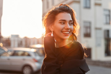 Portrait of a happy pretty young attractive girl with a beautiful smile in stylish black clothes on the background of bright orange sunlight outdoors. Positive girl model walks and enjoys the sunset.