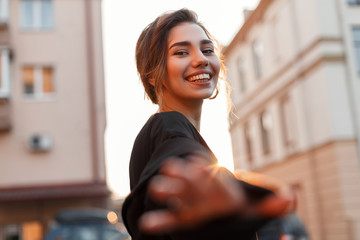 Wall Mural - Positive pretty young woman with a stylish hairstyle with a smile reaching her hand to the camera on the background of a bright orange sunset. Cheerful happy girl walks through the city. Sunny day.