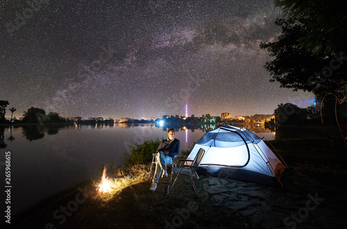 Night Camping On Lake Shore Near Bonfire Man Sitting On