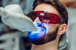 Man having teeth whitening by dental UV whitening device,dental assistant taking care of patient,eyes protected with glasses. Whitening treatment with light, laser, fluoride.