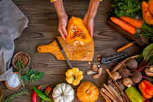 Female Hands Cutting Pumkin On Dark Wooden Kitchen Table With Vegetables Cooking Ingredients, Spoon And Tools, Top View.