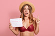 © sementsova321 - Indoor shot of shocked caucasian young female has surprised facial expression, points up with index finger, wearing in red swimsuit and straw hat, holds period calendar, forget date of periods.