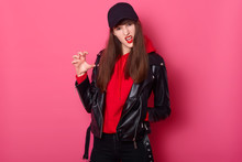 Fashion Young Teen Girl Uses Bright Red Lipstick, Wearing Stylish Hoody, Leather Jacket And Black Cap. Cheeky Tenager Poses Like Tiger. Model Poses In Studio, Looks At Camera Against Pink Wall.