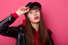 Indoor Shot Of Happy Caucasian Young Teenager Girl In Red Hoodie, Leather Jacket, Stands With Cheeky Facial Expression, Keeps Hand On Black Visor Of Cap Against Rose Studio Wall. People Concept