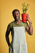 © alfa27 - Happy mid-aged gardener in apron holding potted green plant smiling at camera isolated on yellow