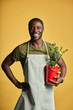 © alfa27 - Happy mid-aged gardener in apron holding potted green plant smiling at camera isolated on yellow