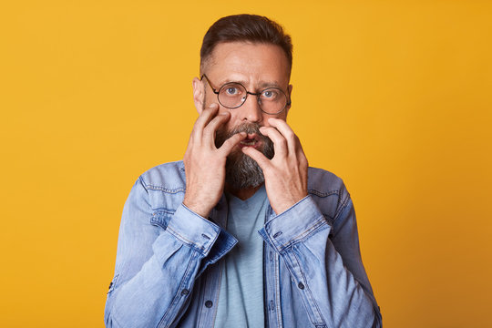 Scared man in panic, dressed in denim jacket, gray shirt and spectacles, feels fear, bits her nails, male watches horror film, model posing isolated over yellow studio background. Human emotions.