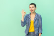 © khosrork - Portrait of funny handsome beautiful short hair young stylish woman in casual striped suit standing with Ok sign and looking at camera with toothy smile. studio shot isolated on light green background