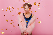 Smiling Curly Girl In Trendy Coat Arranged A Holiday Surprise For Friend's Birthday Party. Laughing Amazing Young Woman Gladly Posing With Golden Glitter Confetti Standing On Pink Background