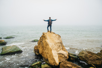 Wall Mural - Traveler with a backpack stands on a rock against a beautiful sea with waves, a stylish hipster boy posing near a calm ocean during a wonderful journey around the world. Shoot from the back