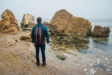 Wall Mural - Traveler with a backpack stands on a rock against a beautiful sea with waves, a stylish hipster boy posing near a calm ocean during a wonderful journey around the world. Shoot from the back