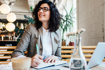 Smiling pretty woman in glasses writing in notepad with pen, sitting in city cafe with coffee and laptop