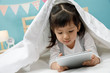 © DN6 - Child girl playing tablet in the blanket on the wooden bed in her bedroom, Happy asian child little girl using computer, learning concept at home