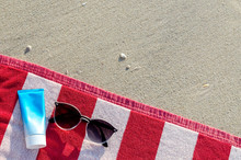 Sunglasses With Sunscreen Lotion And Bag On Red Towel Put On Sand For A Trip On Summer Season At The Beach.