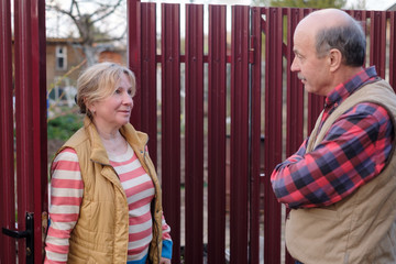 Two senior neighbors takling to each other on sunny day near fence.