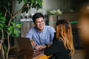 An attractive looking Asian man conversing with a colleague inside a bistro. He is solid in his blue polo.