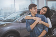 © Ihor - Happy loving young couple smiling at each other, embracing after buying new automobile at car dealership. Beautiful woman hugging her boyfriend from behind, cuddling, celebrating renting modern car