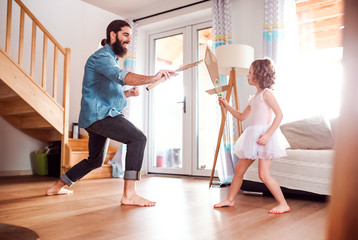 Wall Mural - A small girl and young father with paper swords at home, playing.